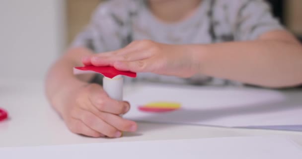 Zbliżenie do dłoni małych chłopców, którzy nakładają klej na pocięty z papieru przedmiot, podczas wykonywania aplikacji z kolorowego papieru i kleju, siedząc przy stole w kuchni — Wideo stockowe