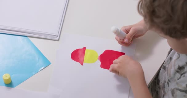 Закрывают руками маленьких мальчишек, которые накладывают жвачку на вырезанные из бумаги предметы, делают аппликации цветной бумагой и жвачкой, сидя за столом на кухне — стоковое видео