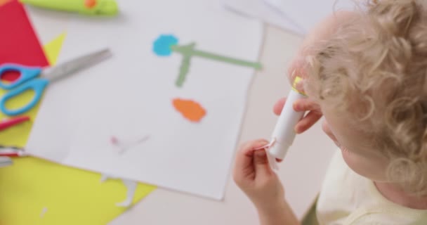 Крупный план маленьких девочек руки, которые кладет клей на вырезанные из бумаги объекты, во время выполнения приложений с цветной бумагой и клеем, сидя за столом на кухне — стоковое видео