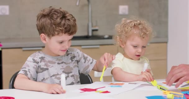 小さな女の子と小さな男の子は、着色された紙と接着剤でアプリケーションを作っています,そして彼らの父は彼らが台所のテーブルに座っているのを助けます — ストック動画