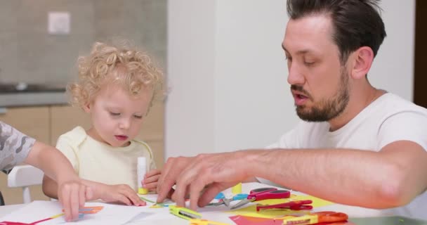 Father with beard and mustaches helps small girl to make an applications with the coloured paper and glue They are sitting at the kitchen table Coloured paper and scissors are at the table in front of — Stock Video