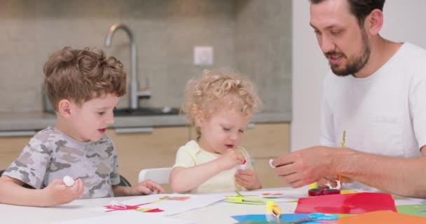 Küçük bir kız ve küçük bir oğlan renkli kağıt ve yapıştırıcıyla başvuru yapıyorlar ve babaları onlara yardım ediyor. Mutfak masasında oturup konuşuyorlar. — Stok video