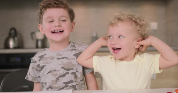 Adorable hermano y hermana sonríen y ríen juntos mientras están sentados en la mesa de la cocina — Vídeos de Stock