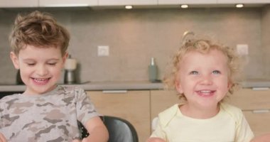 Sevimli erkek ve kız kardeş birlikte gülümseyip gülerken mutfak masasında oturan çocuk elindeki kağıt parçalarından bazılarını tutuyor.