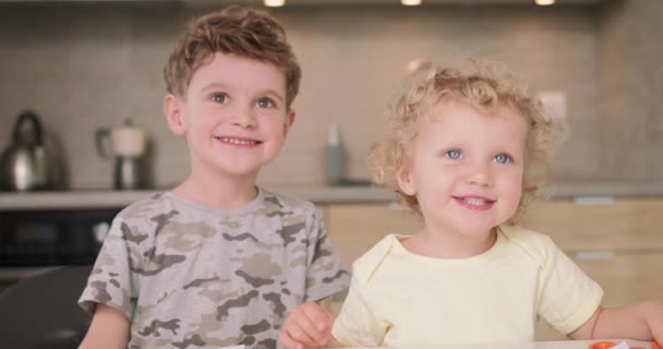 Adorable brother and sister smile and laugh together while sitting at the kitchen table Boy puts his hands on her head, then they high-five — Stock Video