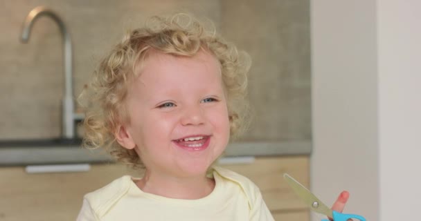 Adorable little girl smiles and laughes while sitting at the kitchen table She has a scissors in her hands — Stock Video