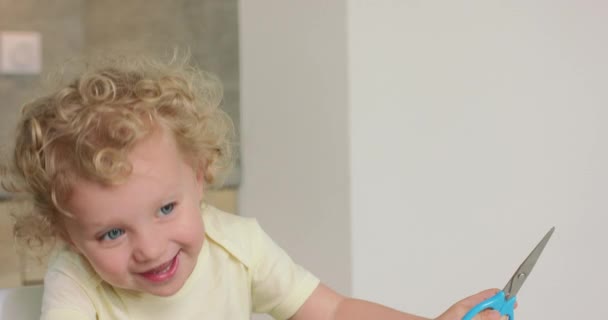 Adorable little girl smiles and laughes while sitting at the kitchen table She has a scissors in her hands — Stock Video