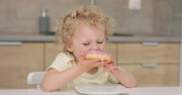 Чарівна маленька дівчинка бере пончик з тарілки, кусає і кладе його назад до тарілки, сидячи за кухонним столом — стокове відео