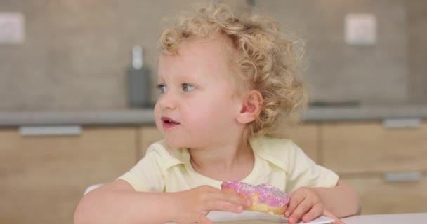 Urocza dziewczynka patrzy w lewo, potem bierze pączka z talerza i gryzie, siedząc przy kuchennym stole. — Wideo stockowe