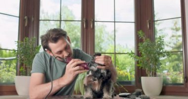 Sakallı ve bıyıklı adam kırpıcı köpeği olan Yorkshire teriyeri masanda oturuyor, makası ve eklentilerin olduğu yerde ev bitkileri ve arka planda bir pencere var.