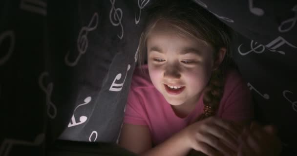 Cara de nad sonriente ondeando con la mano linda adolescente rubia, que utiliza la tableta para charlar, mientras se esconde debajo de la manta en su cama en la noche — Vídeos de Stock