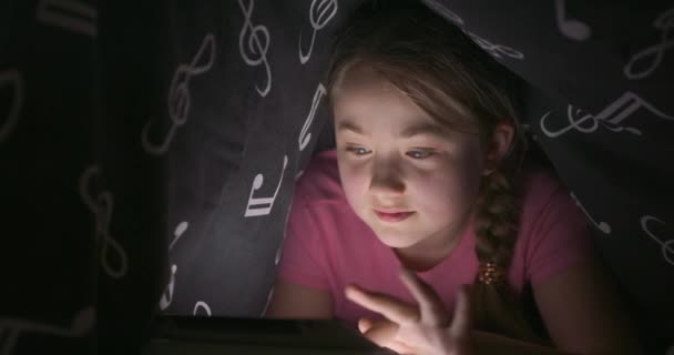 Ansiktet på leende söt tonåring blond flicka med hjälp av tablett medan du gömmer dig under filten i sängen, på natten — Stockvideo
