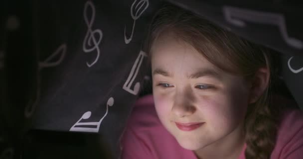 Ansiktet på leende söt tonåring blond flicka med hjälp av tablett medan du gömmer dig under filten i sängen, på natten — Stockvideo