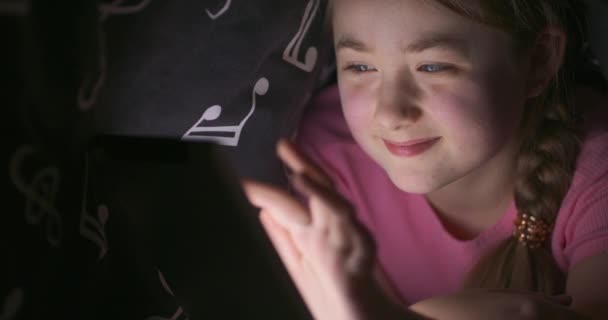 Twarz uśmiechniętej nastoletniej blondynki korzystającej z tabletu ukrywając się pod kocem w łóżku, w nocy — Wideo stockowe