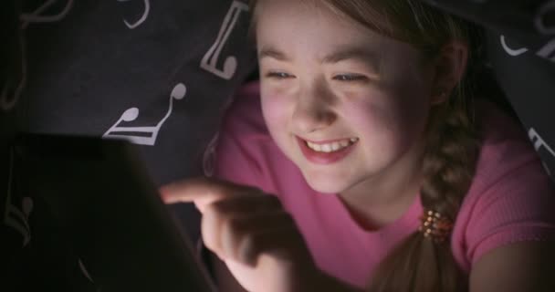 Cara de sonrisa linda adolescente rubia usando tableta mientras se esconde debajo de la manta en su cama, por la noche — Vídeos de Stock