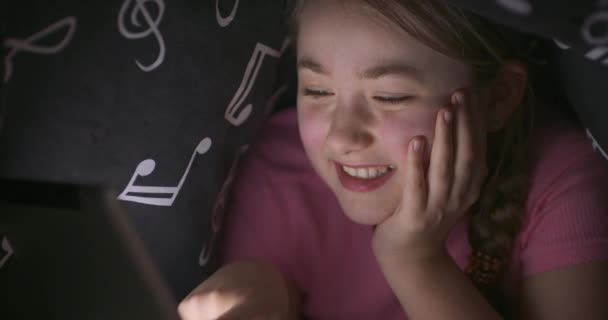 Πρόσωπο του χαμόγελου χαριτωμένο έφηβος ξανθιά κοπέλα χρησιμοποιώντας tablet, ενώ κρύβεται κάτω από την κουβέρτα στο κρεβάτι της, τη νύχτα Στην αρχή υποστηρίζει το κεφάλι της με το χέρι της, αλλά nex βάζει το χέρι προς τα κάτω — Αρχείο Βίντεο