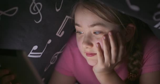 Rosto de menina loira adolescente bonito, que está lendo algo usando tablet enquanto se esconde sob o cobertor em sua cama, à noite, apoiando sua cabeça com a mão — Vídeo de Stock