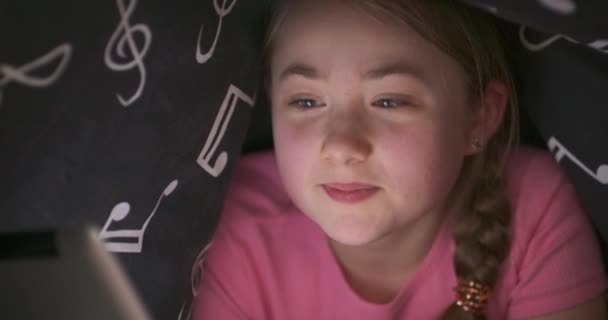 Gezicht van glimlachende nad zwaaien met de hand schattig tiener blond meisje, die gebruikt tablet om te chatten, terwijl is ondergedoken onder de deken in haar bed in de nacht — Stockvideo