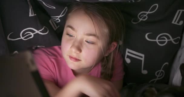Close up of a face of smiling cute teenage blonde girl uses the tablet while hiding under the blanket in her bed, at the night She pets and hugs her yorkshire terrier with her cheek — Stock Video
