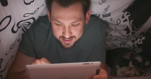 Gros plan du visage d'un jeune homme barbu et moustachu, utilisant une tablette numérique sous la couverture, dans son lit, la nuit Son terrier du yorkshire est couché à côté de lui — Video