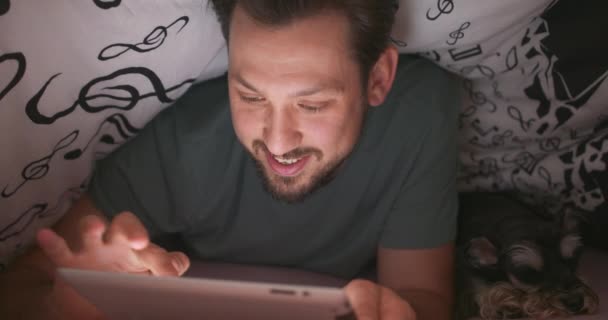 Gros plan du visage souriant d'un jeune homme barbu et moustachu, utilisant une tablette numérique sous la couverture, dans son lit, la nuit. Son terrier du yorkshire se trouve à côté de lui.. — Video