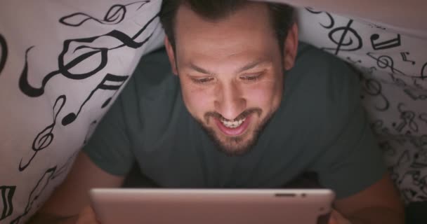 一个留着胡子、满脸胡须、满脸胡须的年轻人的脸靠得很近，晚上在床上用数码平板电脑在毯子下聊天、挥手 — 图库视频影像