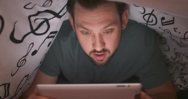 Zbliżenie do twarzy młodego brodatego i muskularnego mężczyzny, używającego tabletu cyfrowego pod kocem, w łóżku, w nocy Jest zaskoczony czymś, co czyta — Wideo stockowe