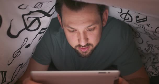 Close-up do rosto de um jovem barbudo e homem de bigode, usando tablet digital sob cobertor, em sua cama, à noite Ele está surpreso com algo que lê — Vídeo de Stock