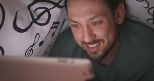Zbliżenie na twarz młodego brodatego i zmutowanego mężczyzny, który macha cyfrowym tabletem podczas gry, pod kocem, w łóżku, w nocy — Wideo stockowe