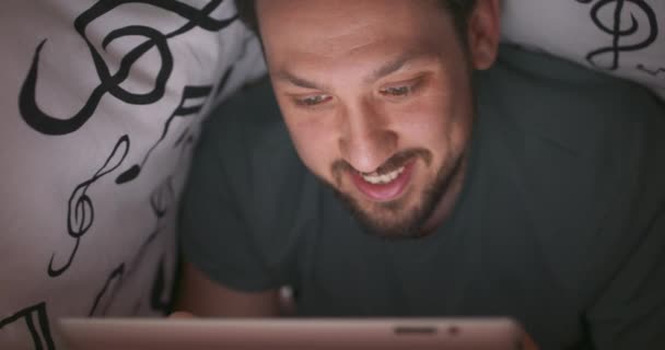 Close-up do rosto de um jovem barbudo e bigodinho, que acena tablet digital durante um jogo, sob cobertor, em sua cama, à noite — Vídeo de Stock