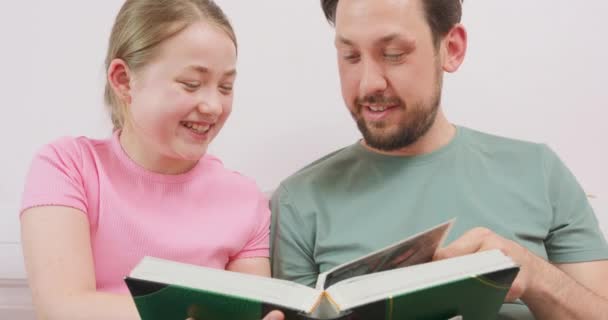 Close up of father and laughing teen daughter are sitting on the sofa, and are looking at the photo album — Stock Video