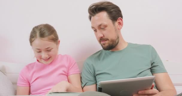 Blisko ojca, trzymając tabliczkę i śmiejąc się nastolatka córka siedzi na kanapie, i patrząc na album fotograficzny — Wideo stockowe