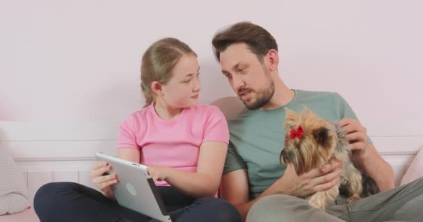 Father, with a dog on his palm, and teen daughter, holding a tablet, are sitting on the sofa in living room, browsing the online store, enjoying weekend Father agrees to choose the thing, his daughter — Stock Video