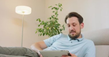 Genç sakallı adam kanepede uzanıyor ve elinde tableti tutuyor. Tableti gevşetiyor ve kaydırıyor. Arka planda beyaz duvarlar, lamba ve bir bitkinin yeşil yaprakları var.