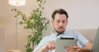 Genç sakallı adam kanepede uzanıyor ve elinde tableti tutuyor. Tableti gevşetiyor ve kaydırıyor. Arka planda beyaz duvarlar, lamba ve bir bitkinin yeşil yaprakları var.