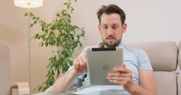 Νεαρός γενειοφόρος άνδρας είναι ξαπλωμένος στον καναπέ και κρατώντας tablet στα χέρια Είναι χαλαρωτικό και παίζει ένα παιχνίδι στο tablet Λευκό τοίχους, λάμπα και ένα πράσινο φύλλα ενός φυτού στο παρασκήνιο — Αρχείο Βίντεο