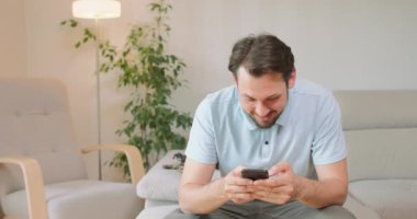 Genç sakallı adam kanepede oturuyor, akıllı telefon kullanıyor, muhtemelen bir oyun oynuyor ya da bir şeyler yazıyor ve arka planda beyaz duvarlar, lamba ve yeşil yapraklar var.