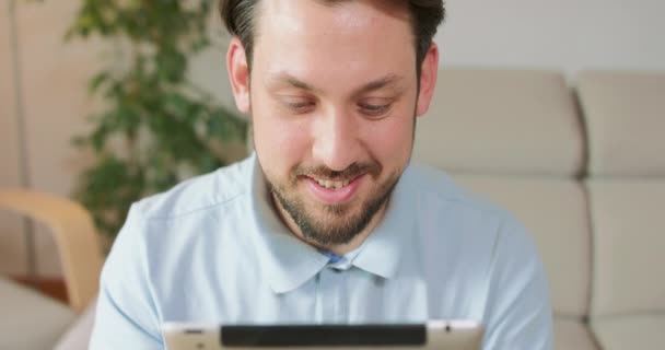 Closup of young bearded mans face while he is holding and using tablet, probably playing a game or typing something White walls and a green leaves of a plant in the background — Stock Video