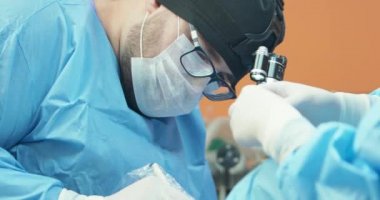 Erkek veteriner, gözlüklü, beyaz eldivenli, kepli ve cerrahi kıyafetli, ameliyat sırasında anestezi edilmiş bir köpeğin arka bacağını ameliyat ederken.