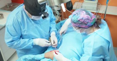 En iyi kamera, erkek veteriner, gözlüklü, beyaz eldivenli, kepli ve cerrahi kıyafetli, ameliyat edilen köpeğe dikiş atarken cerrahi iplikli bir kadın asistanı ona yardım ederken.