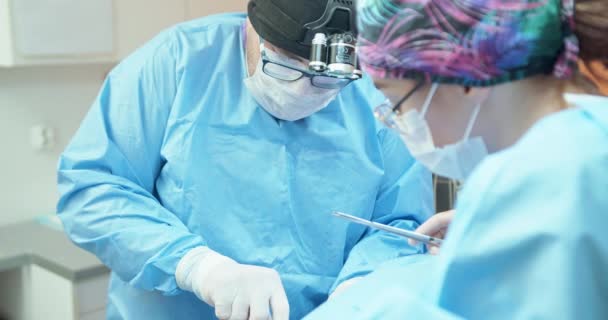 Veterinario masculino, en gafas, guantes blancos, gorra y en traje quirúrgico, mientras sutura la pierna de los perros operados con un hilo quirúrgico Ayudante femenina le ayudan sosteniendo la cortina quirúrgica y — Vídeos de Stock