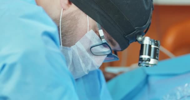 Κοντινό πλάνο πρόσωπο του αρσενικού κτηνιάτρου, σε γυαλιά, λευκά γάντια, καπέλο και σε χειρουργική στολή, ενώ ο ίδιος λειτουργεί το πίσω πόδι ενός αναισθητοποιημένου σκύλου κατά τη διάρκεια της χειρουργικής επέμβασης Στο παρασκήνιο - πορτοκαλί τοίχοι — Αρχείο Βίντεο