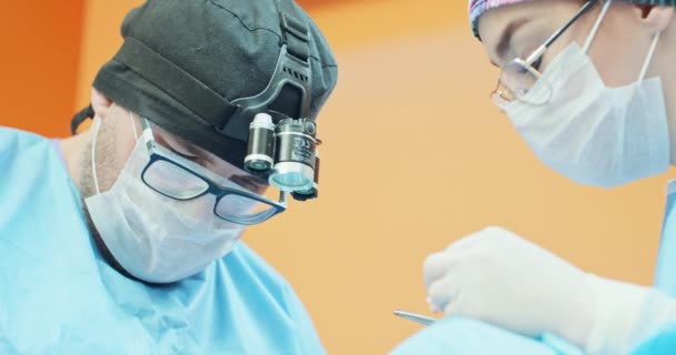 Visage rapproché du vétérinaire masculin, en lunettes, casquette et tenue chirurgicale, alors qu'il opère la jambe arrière d'un chien anesthésié pendant la chirurgie Un assistant féminin légèrement flou l'assiste par — Video