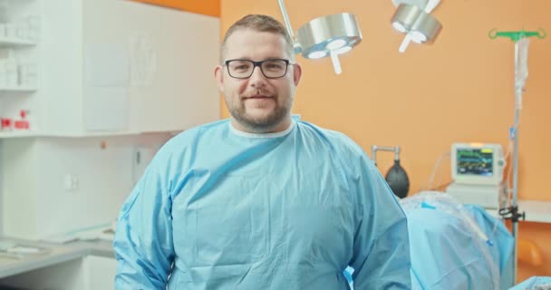 Homme vétérinaire, à lunettes, en tenue chirurgicale, croise les bras sur sa poitrine et sourit, après l'intervention chirurgicale sur la jambe des chiens En arrière-plan - murs orange flous, recouverts de chirurgie — Video