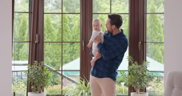 Padre barbudo y bigotudo sostiene a su adorable bebé y se queda con él delante de la ventana, besando la ventana del bebé y una pared blanca en el fondo — Vídeo de stock