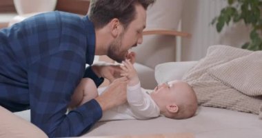 Sakallı ve bıyıklı baba, bebeğin yanında diz çöküyor, göğsünü okşuyor, gülümsüyor. Bebek, babasının çenesine, sandalyesine, kanepesine, yeşil bitkilere ve beyaz bir duvara dokunuyor.