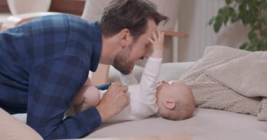 Sakallı ve bıyıklı baba bebeğin yanında diz çöküyor, onu gıdıklıyor, gülümsüyor bebeğin babasının çenesine, yüzüne ve kafasına dokunuyor. Elleriyle sandalye, kanepe, yeşil bitkiler ve beyaz bir duvara.