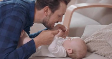 Sakallı ve bıyıklı baba bebeğin yanında diz çöküyor, elleriyle oynuyor, parmaklarıyla dokunuyor yanaklarıyla, boynuyla, göğsüyle ve burnu bebeğin karnına değiyor.