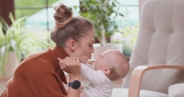 Anneye yakın çekim, bebek kanepede sırtüstü yatarken dizlerinin üzerine çöker Anne bebeği biraz alır ve nazikçe öper Koltuk, dalak bitkileri ve arka planda bir pencere
