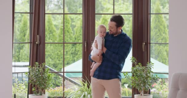 Бородатый и усатый отец держит своего очаровательного ребенка и слегка машет ногами, улыбаясь перед окном и белой стеной на заднем плане. — стоковое видео